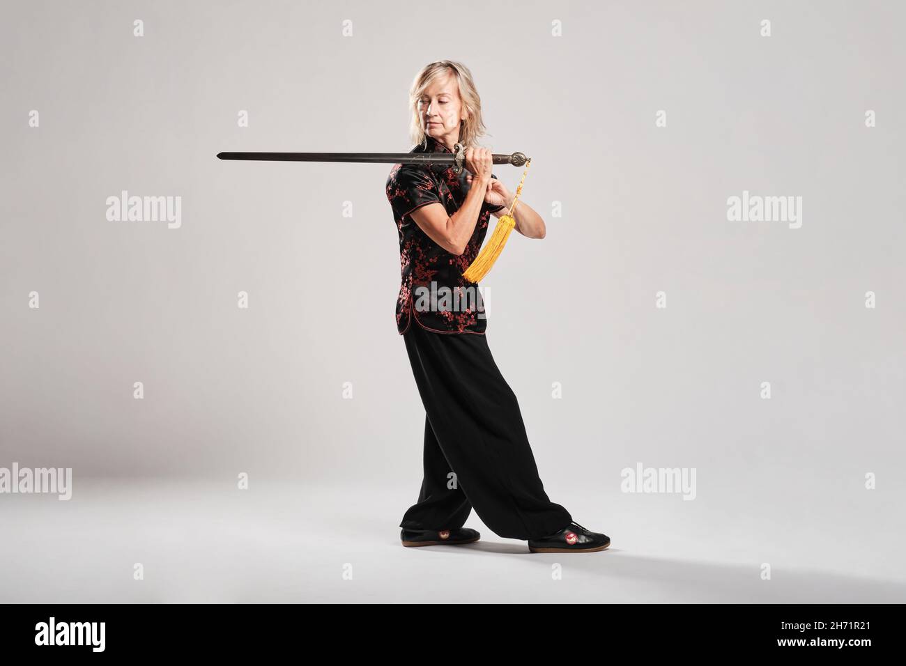 Femme blonde mûre pratiquant le Tai Chin avec une épée dans un fond blanc portant une veste chinoise traditionnelle noire avec une décoration chinoise rouge, noire Banque D'Images