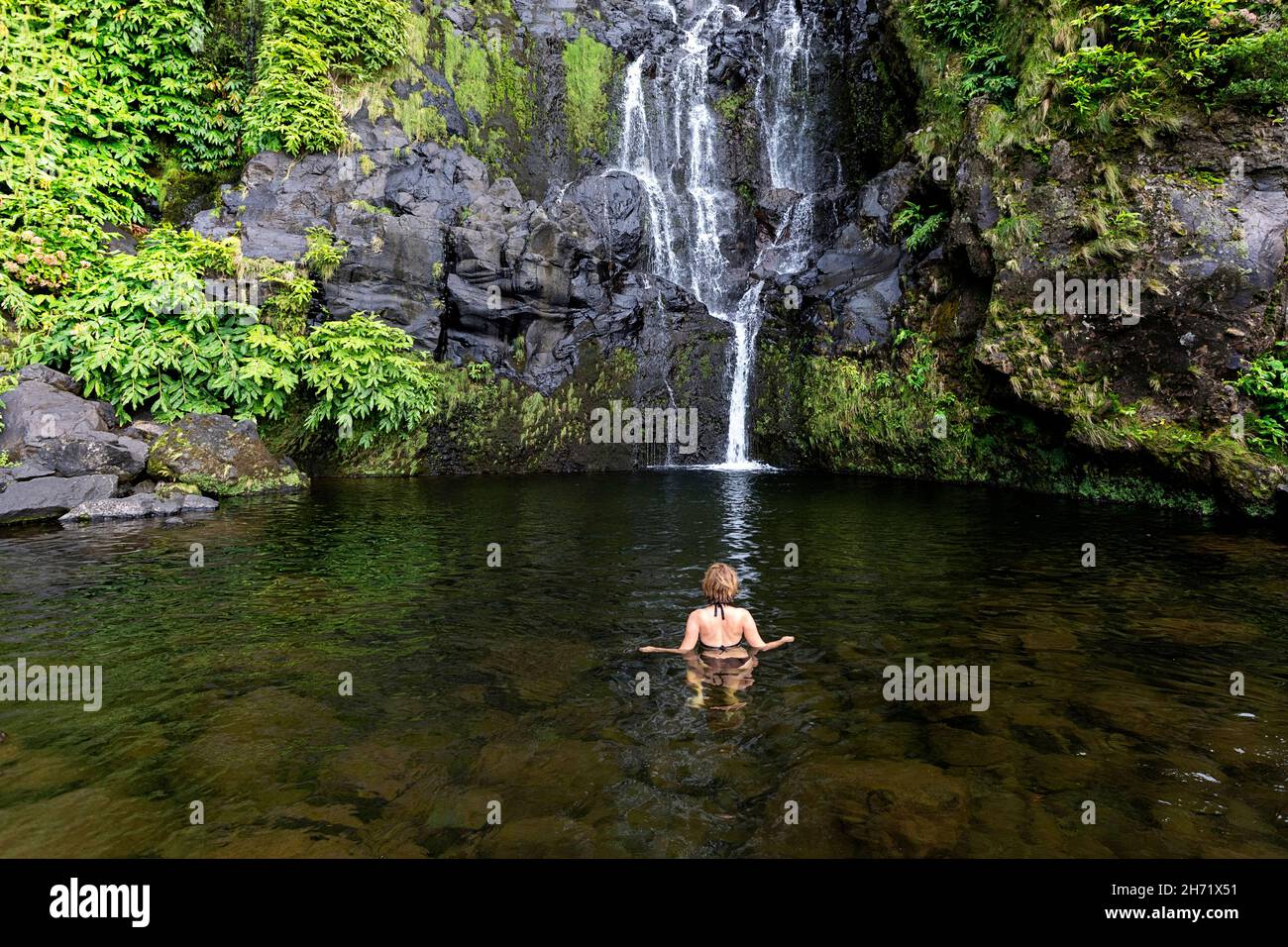 Femme se baignant sous une douche en cascade dans la piscine naturelle de Cascata do Poco do Bacalhau cascade à Faja Grande, Banque D'Images