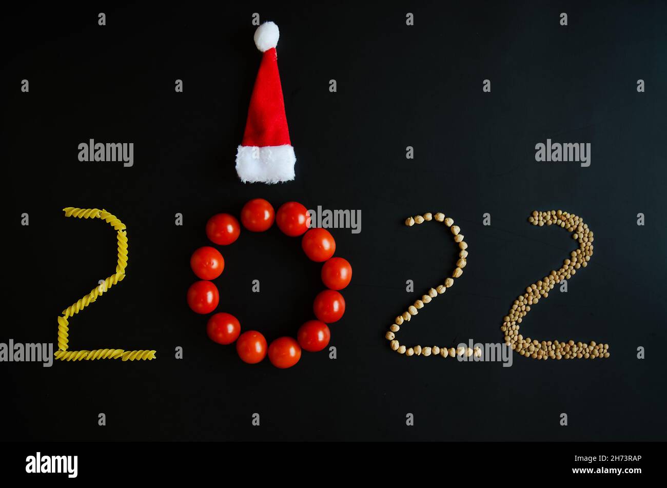 Numéro de la nouvelle année 2022. Symbole de la nouvelle année.Concept créatif vacances.Aliments végétariens sains. Tomates rouges, pâtes, chapeaux de Noël sur fond noir. Banque D'Images