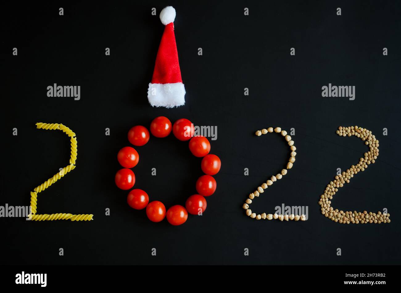 Numéro de la nouvelle année 2022. Symbole de la nouvelle année.Concept créatif vacances.Aliments végétariens sains. Tomates rouges, pâtes, chapeaux de Noël sur fond noir. Banque D'Images