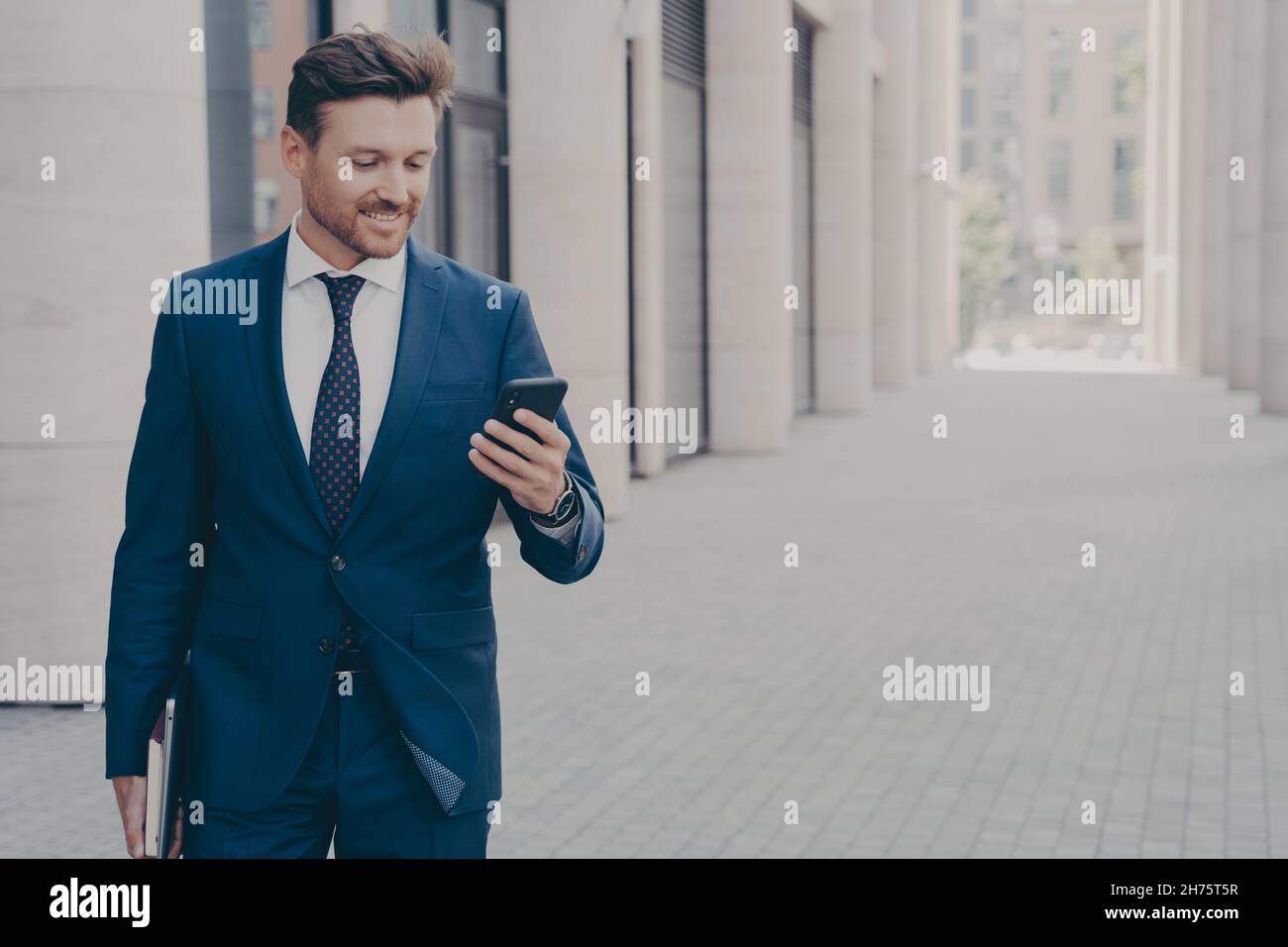 Jeune homme souriant Président-directeur général réussi regardant un smartphone avec une expression satisfaite à l'extérieur Banque D'Images