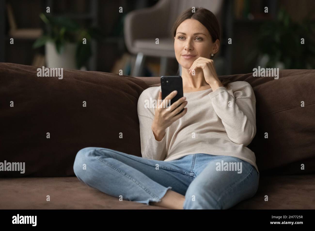 Une jeune femme s'assoit sur un canapé pour tenir un smartphone qui a l'air pensif Banque D'Images