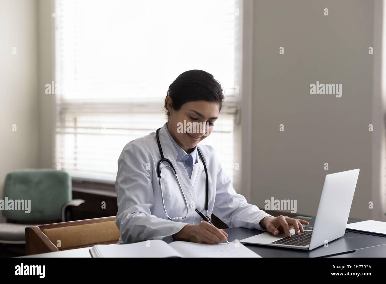 Femme indienne souriante médecin prenant des notes, en utilisant un ordinateur portable dans le bureau Banque D'Images