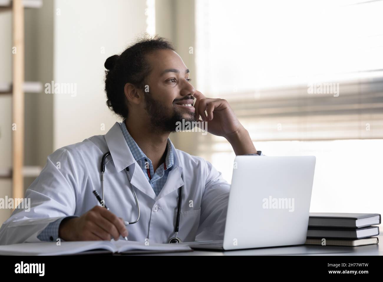 Homme afro-américain pensif souriant médecin prenant des notes, à l'aide d'un ordinateur portable Banque D'Images