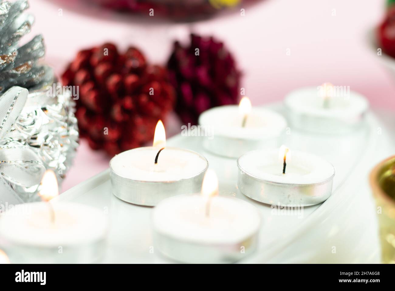 Bougies chauffe-plat blanches illuminées en rangée avec des décorations à paillettes en forme de cône de pin marron sur fond rose clair.Thème pour Joyeux Noël et Joyeux Nouveau Banque D'Images