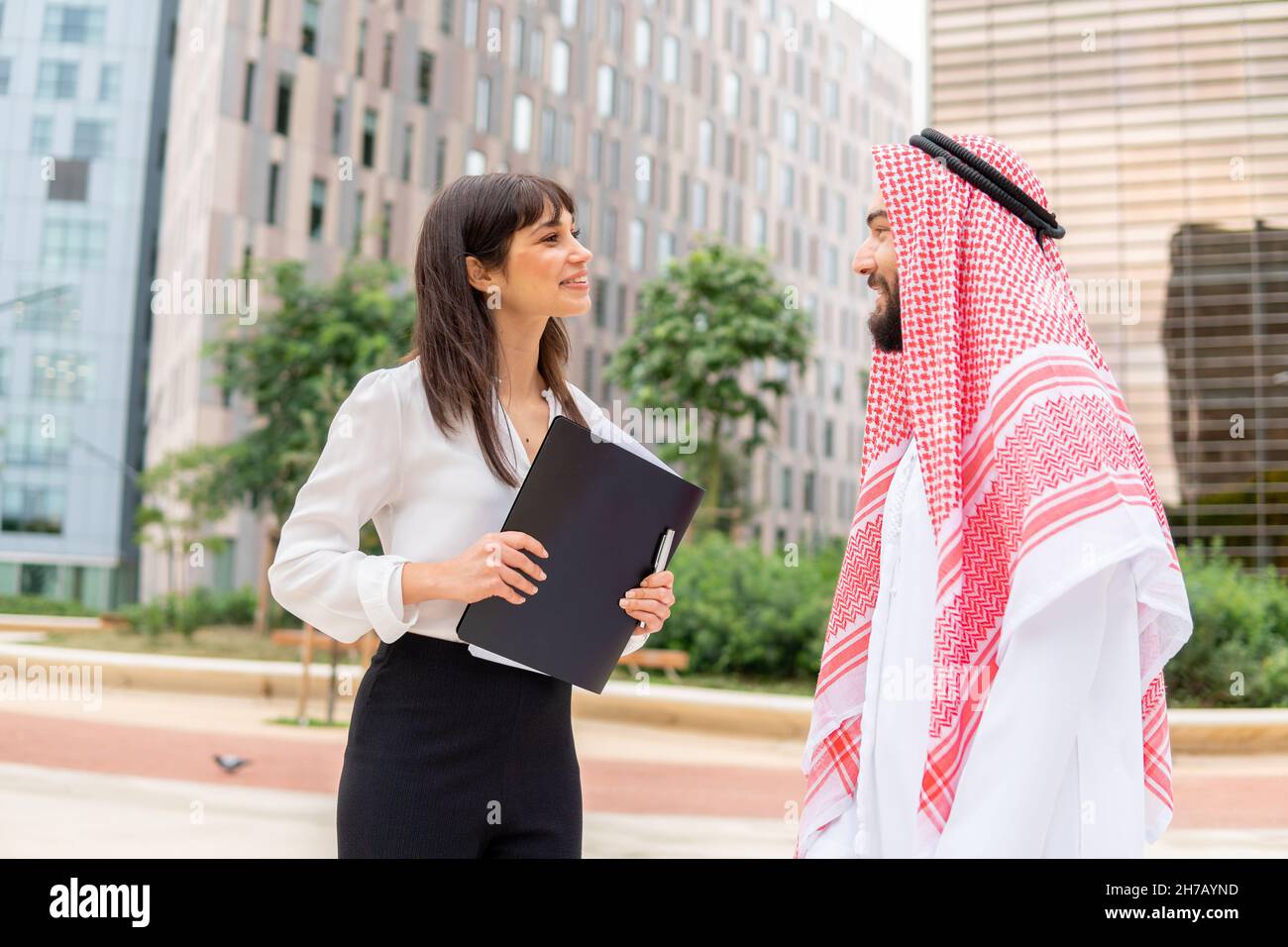 Homme d'affaires arabe satisfait et sa jeune belle assistante avec presse-papiers debout dans la rue de la ville et parlant, homme saoudien discutant de contrat avec une femme d'affaires européenne lors d'une réunion en plein air Banque D'Images