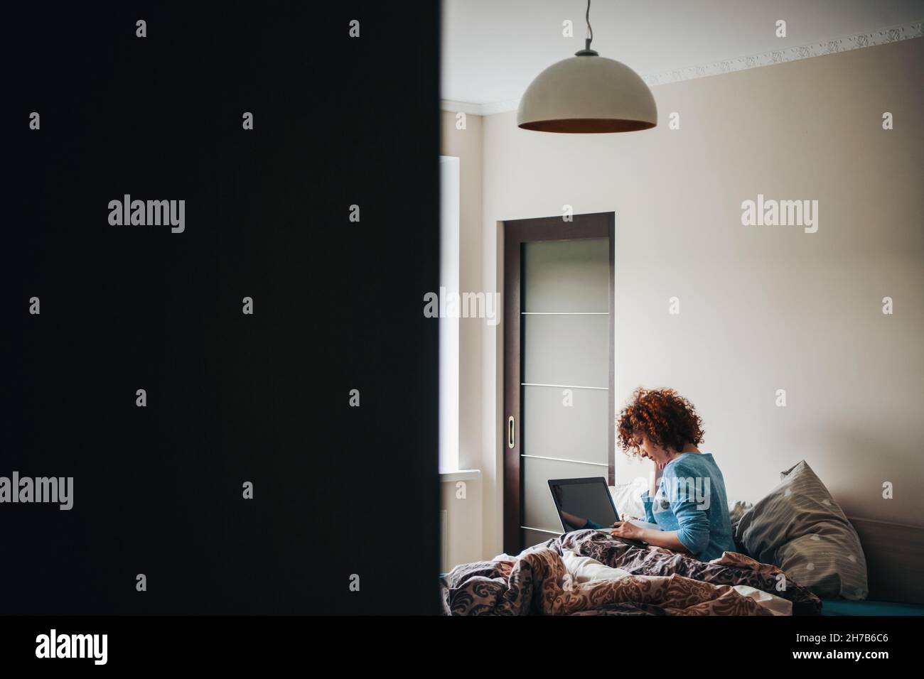 Vue arrière de la femme caucasienne utilisant un ordinateur portable assis sur le lit à la maison.Heure du matin.Femme de Cury.Vue de la porte à moitié ouverte Banque D'Images