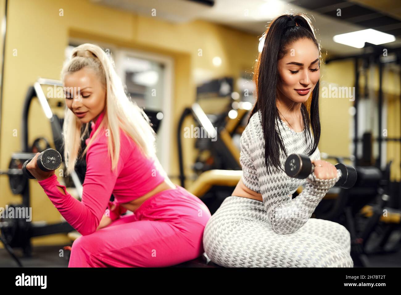 Deux femmes de sport faisant des exercices pour les biceps avec des haltères tout en étant assis sur le banc dans la salle de gym. Banque D'Images