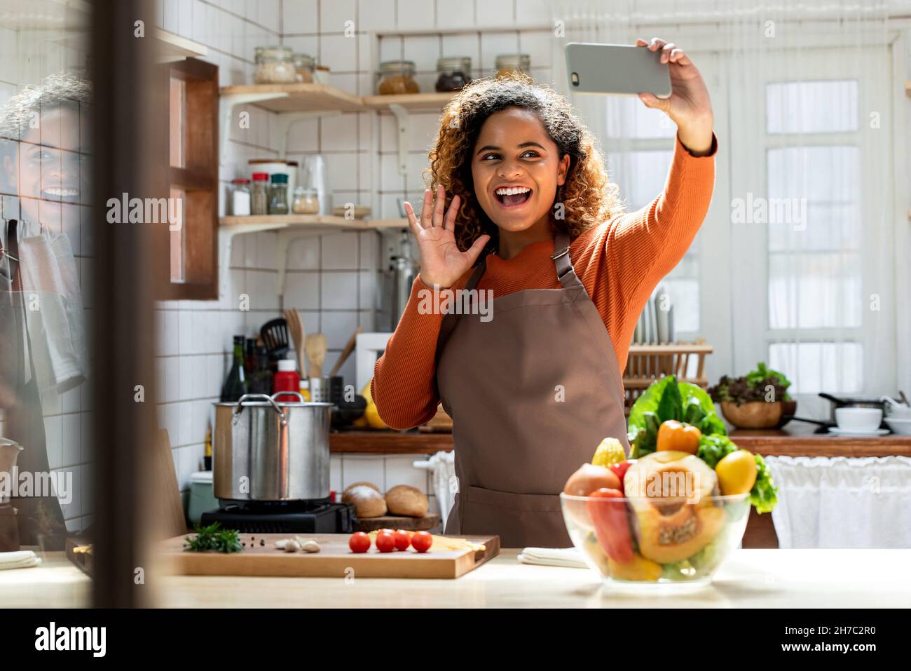 Jeune femme afro-américaine heureuse en train de dire bonjour lors d'un appel vidéo tout en cuisinant dans la cuisine à la maison Banque D'Images