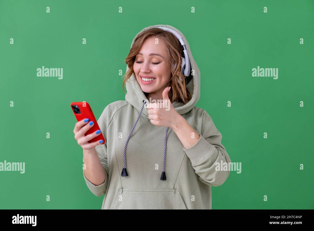 Des dents heureuses sourient femme écouter de la musique casque tenant le smartphone dans Main montrer le pouce vers le haut geste femelle caucasienne apprécier podcast ou livres audio habillés Banque D'Images