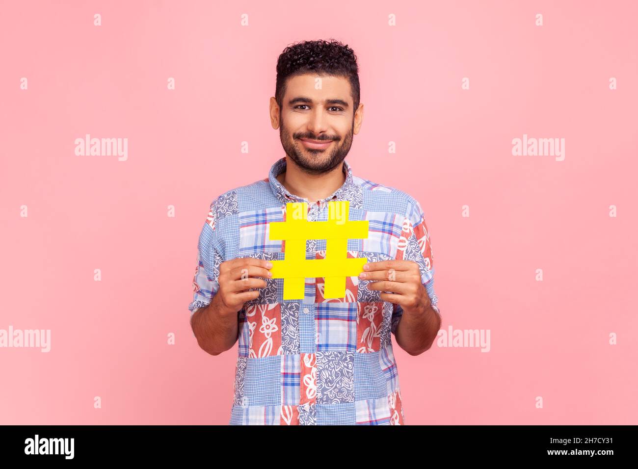 Popularité dans les médias sociaux.Portrait d'un homme heureux aux cheveux foncés avec une barbe dans une chemise décontractée tenant un grand signe de hashtag jaune, regardant l'appareil photo.Studio d'intérieur isolé sur fond rose. Banque D'Images