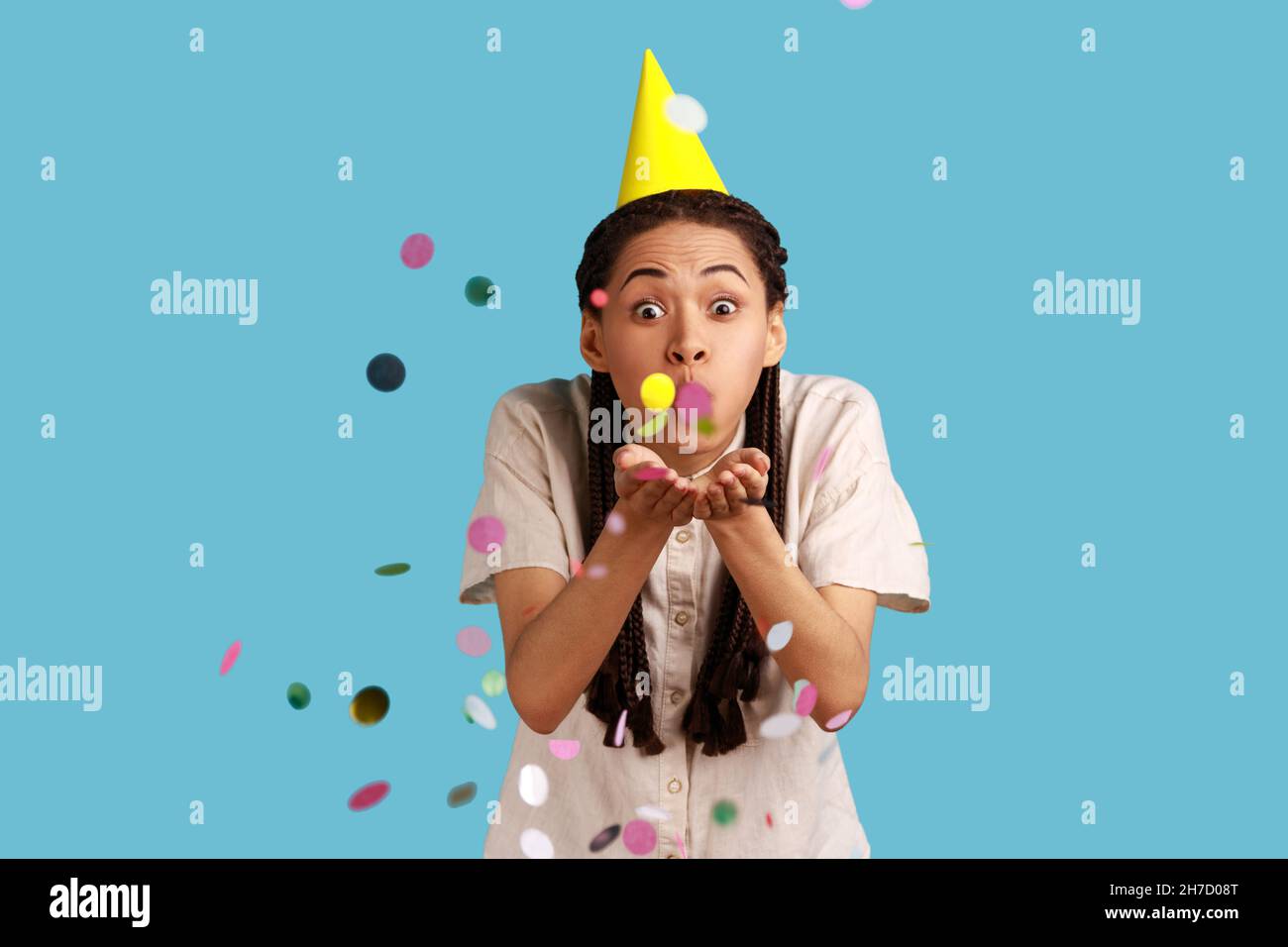 Portrait d'une femme excitée avec des dreadlocks noirs et un cône de fête, des confettis soufflants, célébrant l'anniversaire, l'ambiance festive, portant une chemise blanche.Studio d'intérieur isolé sur fond bleu. Banque D'Images