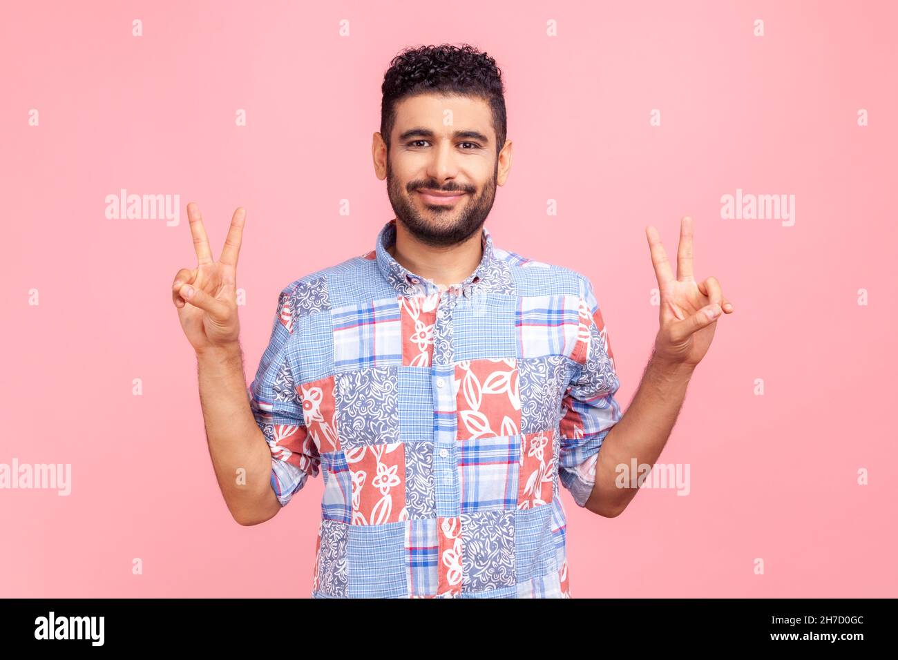 Joyeux homme réussi avec des cheveux foncés et la barbe en bleu chemise décontractée montrant le signe v geste de victoire avec les doigts, regardant l'appareil photo avec le sourire.Studio d'intérieur isolé sur fond rose. Banque D'Images