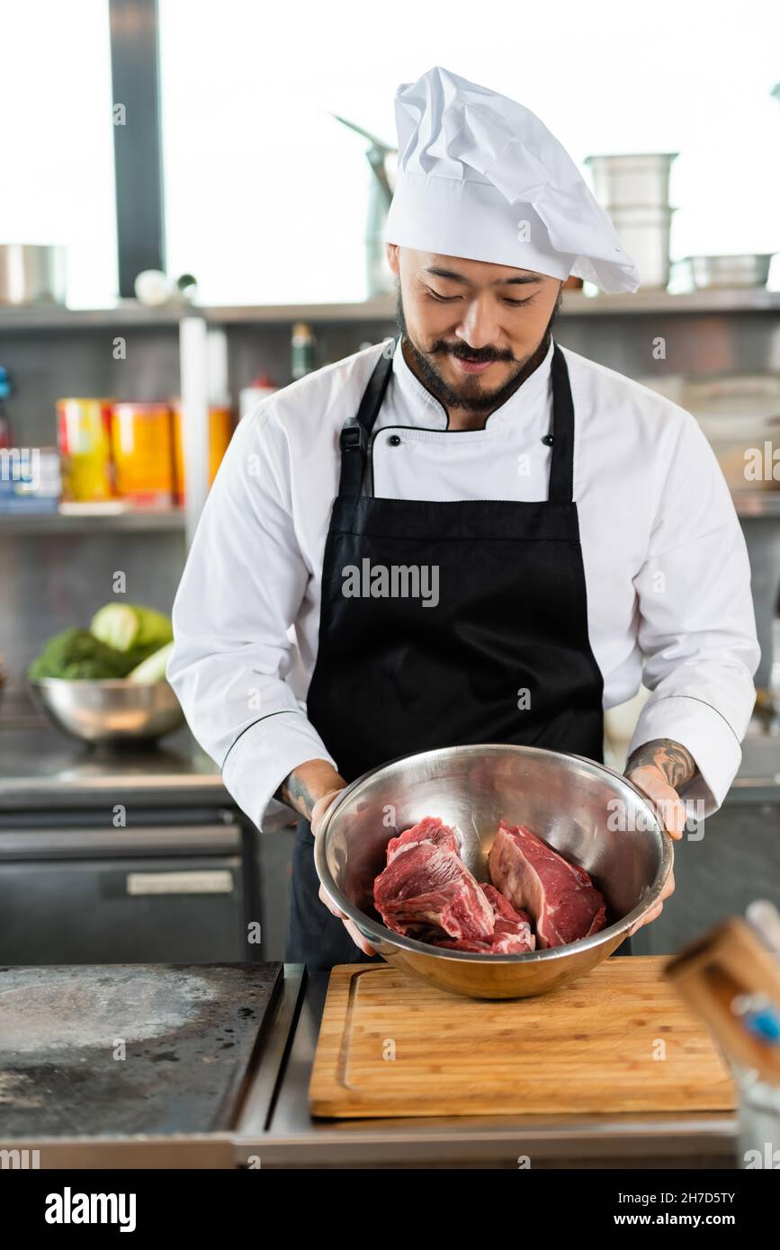 Chef asiatique en tablier et chapeau tenant un bol avec de la viande crue  dans la cuisine Photo Stock - Alamy