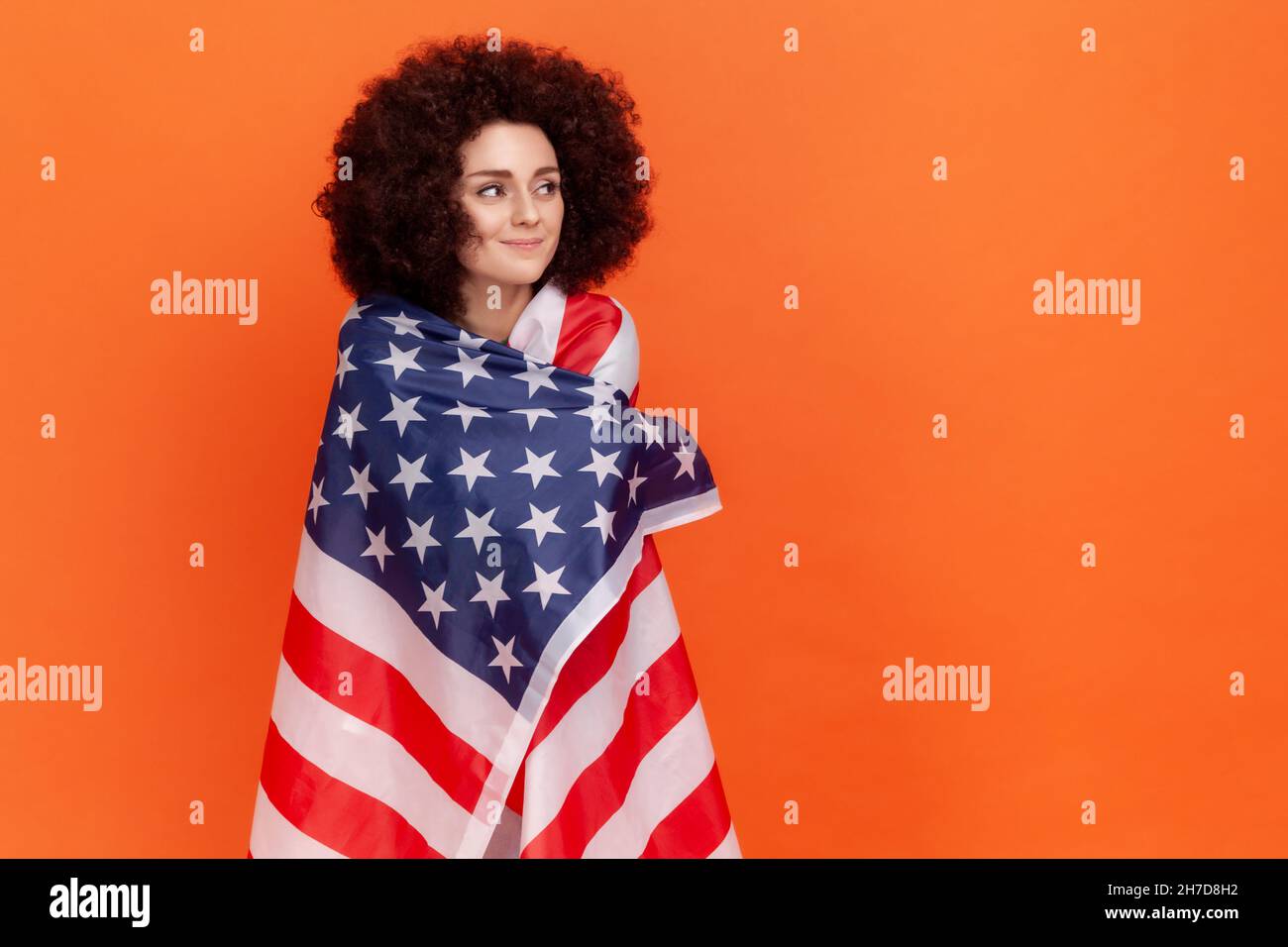 Calme femme rêveuse avec la coiffure afro portant vert décontracté style chandail debout enveloppé dans le drapeau des États-Unis, regardant loin, les droits de l'homme.Studio d'intérieur isolé sur fond orange. Banque D'Images
