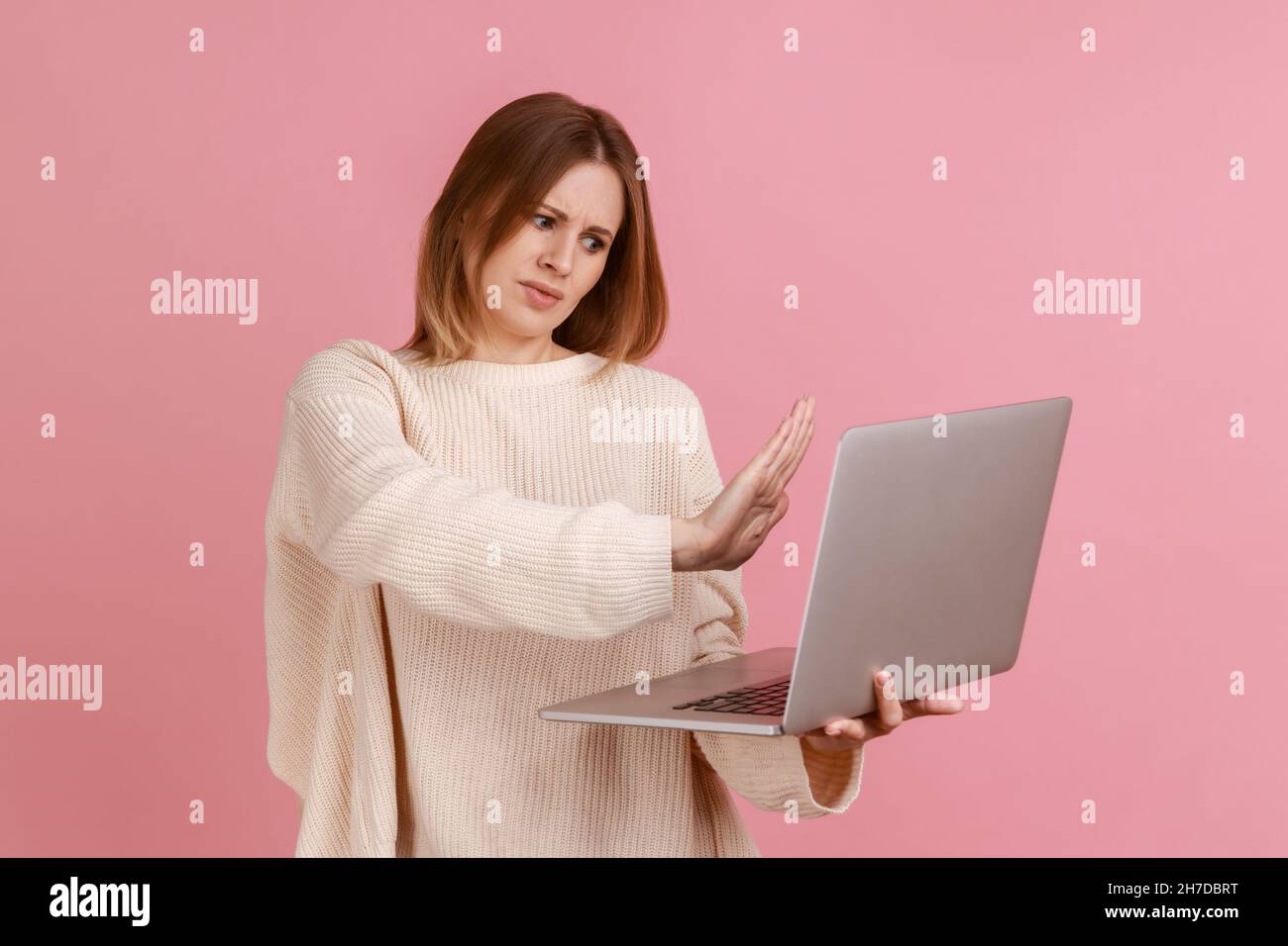 Femme montrant un geste d'arrêt sur l'écran de l'ordinateur portable, avertissement avec un panneau d'interdiction lors d'un appel vidéo, communication en ligne, port d'un chandail blanc.Studio d'intérieur isolé sur fond rose. Banque D'Images