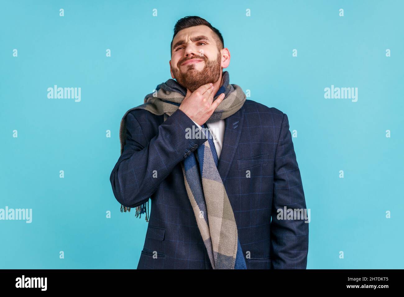 Jeune homme adulte avec barbe portant un costume de style officiel, enveloppé dans un foulard, touchant son cou, souffrant de maux de gorge et de température élevée.Studio d'intérieur isolé sur fond bleu. Banque D'Images