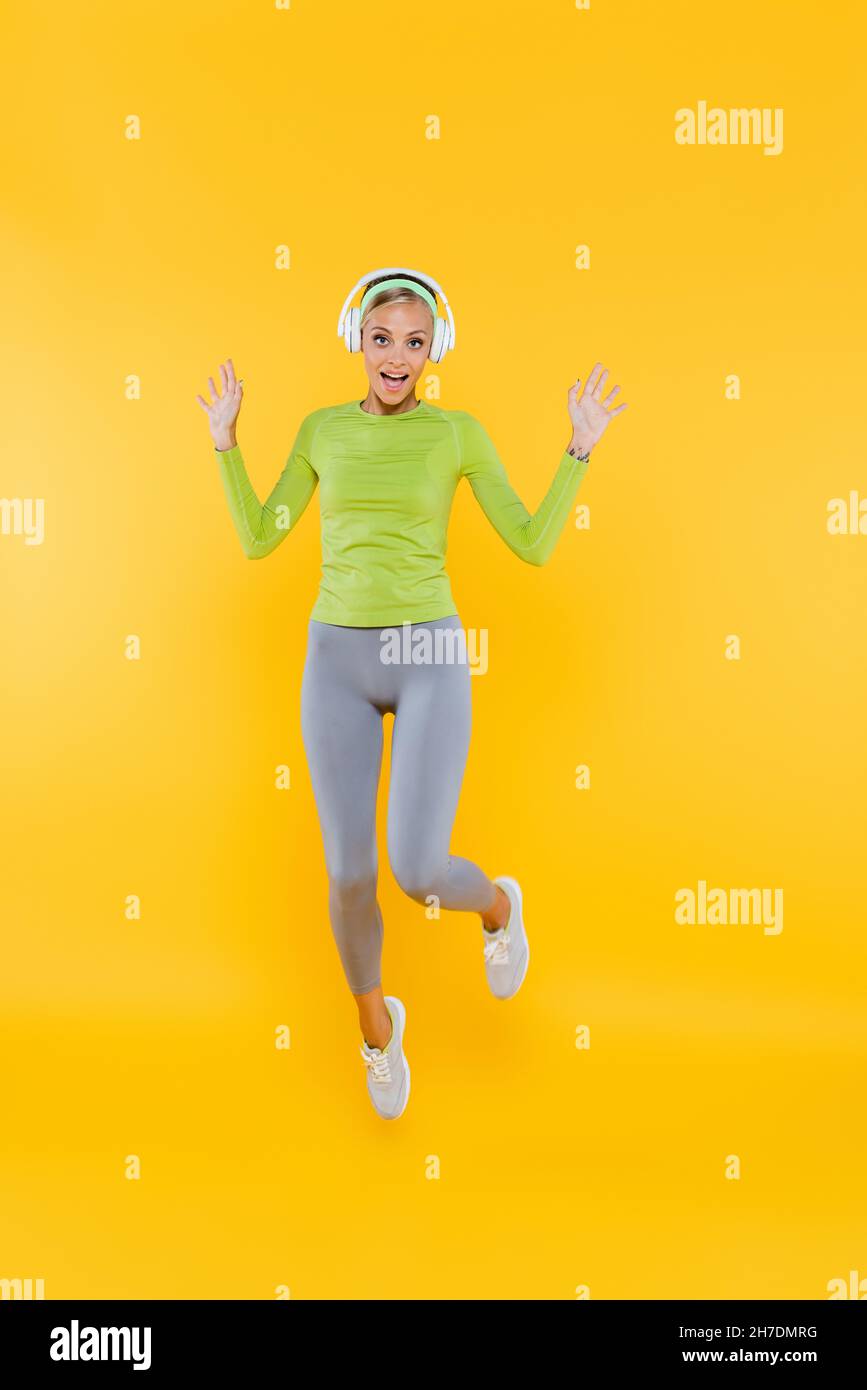 vue intégrale de la femme excitée et sportive dans un casque regardant l'appareil photo tout en sautant sur le jaune Banque D'Images