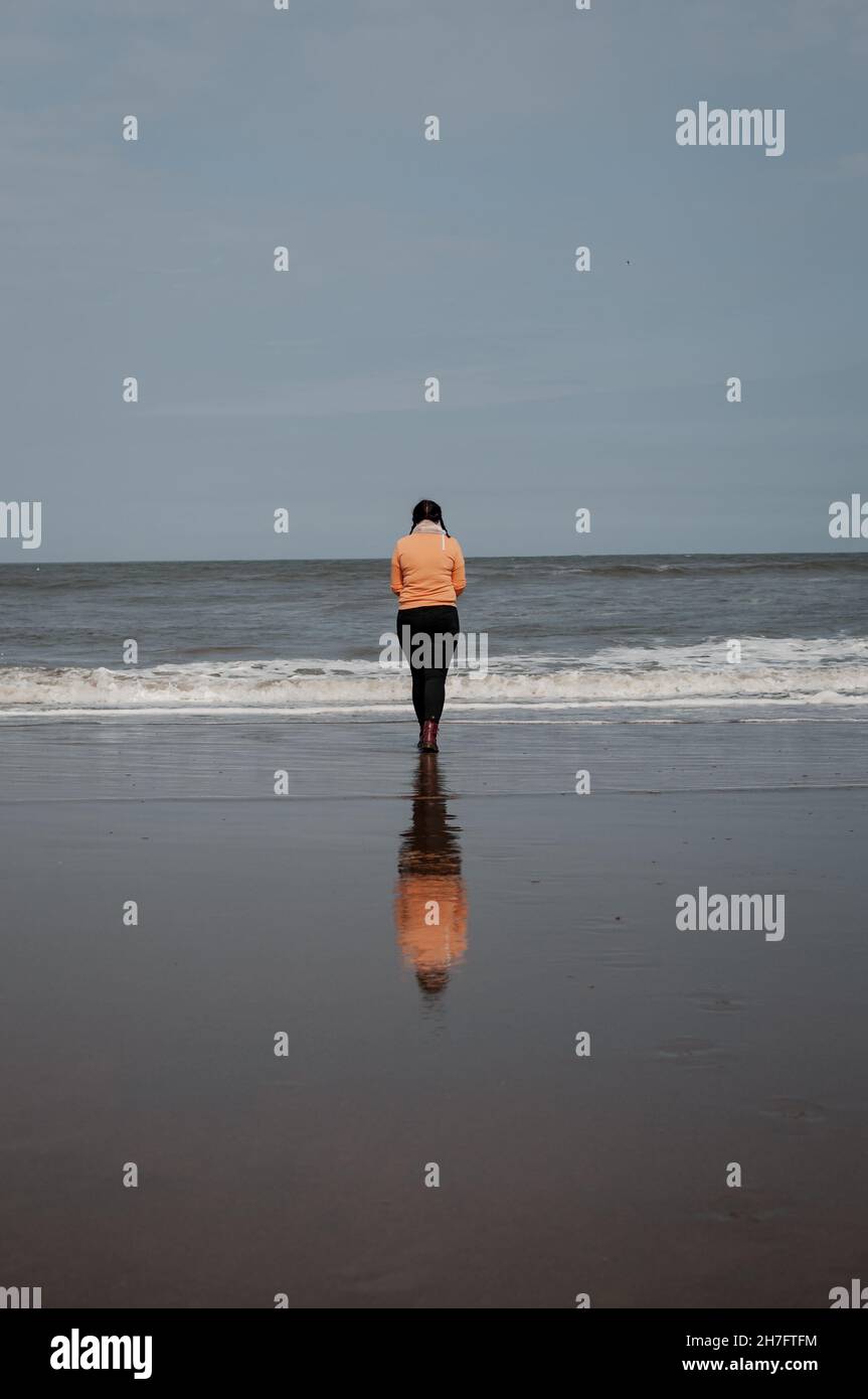 Une figure solitaire se tient au bord de l'océan, sa réflexion se jette sur le sable Banque D'Images