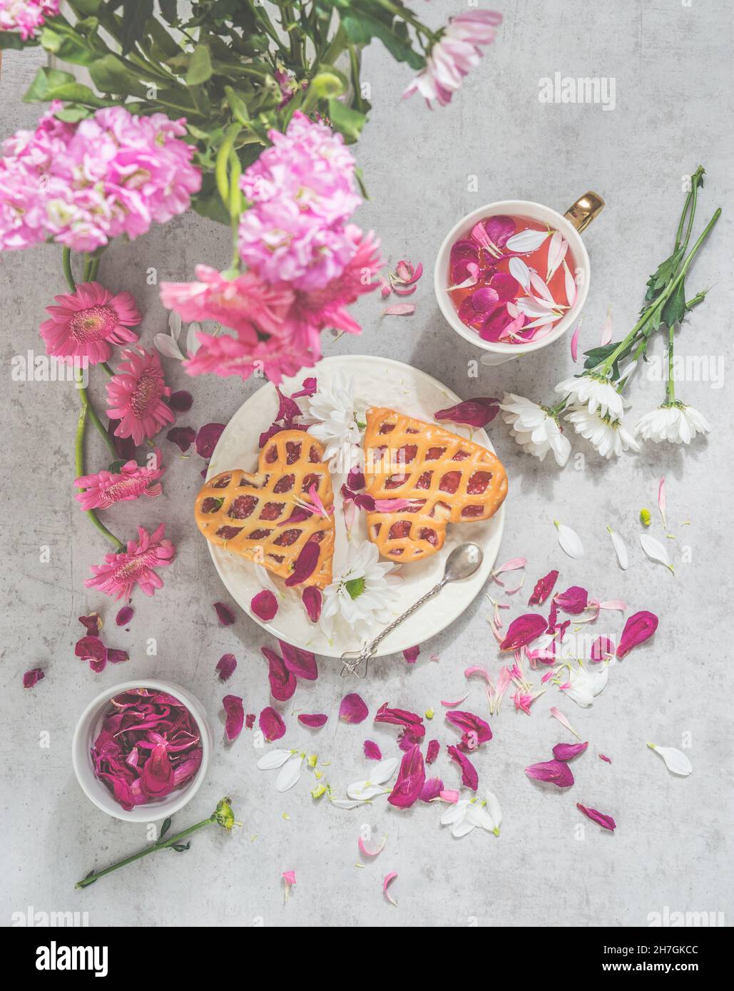 Pause-thé romantique avec thé aux cerisiers en fleurs, pâtisserie en forme de coeur remplie de confiture et de fleurs blanches et roses sur table de cuisine en béton gris.Vue de dessus Banque D'Images