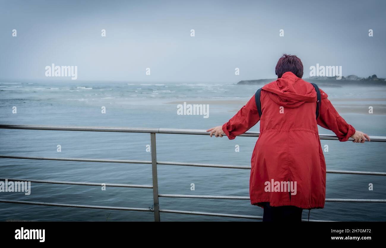Vue arrière d'une femme réfléchissante de trench-coat rouge contemplant la mer sur une plage pendant des vacances d'été, à l'extérieur.Mode de vie moyen. Banque D'Images