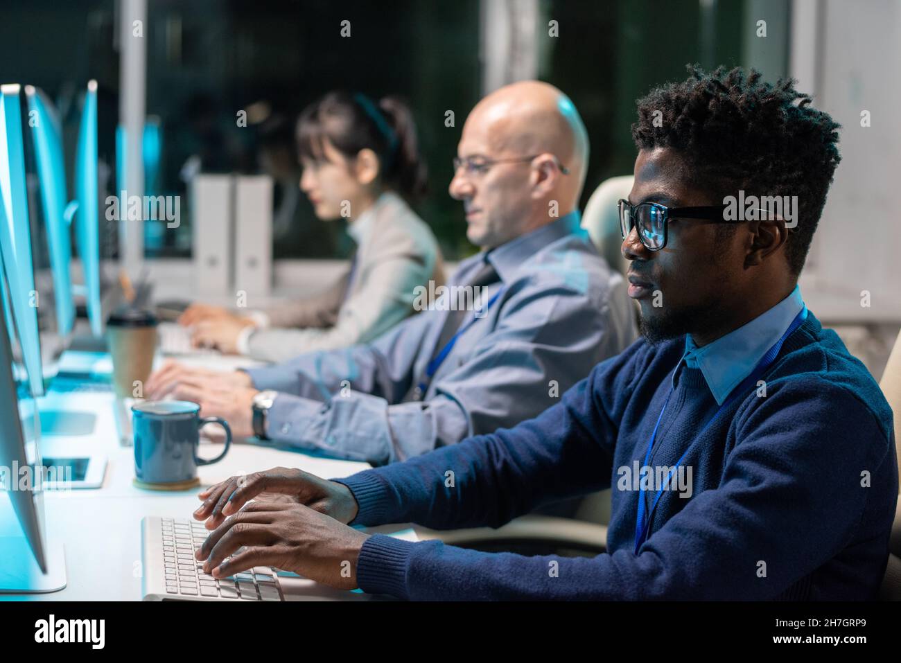 Rangée de programmeurs interculturels contemporains travaillant sur de nouveaux logiciels devant des moniteurs d'ordinateur au bureau Banque D'Images