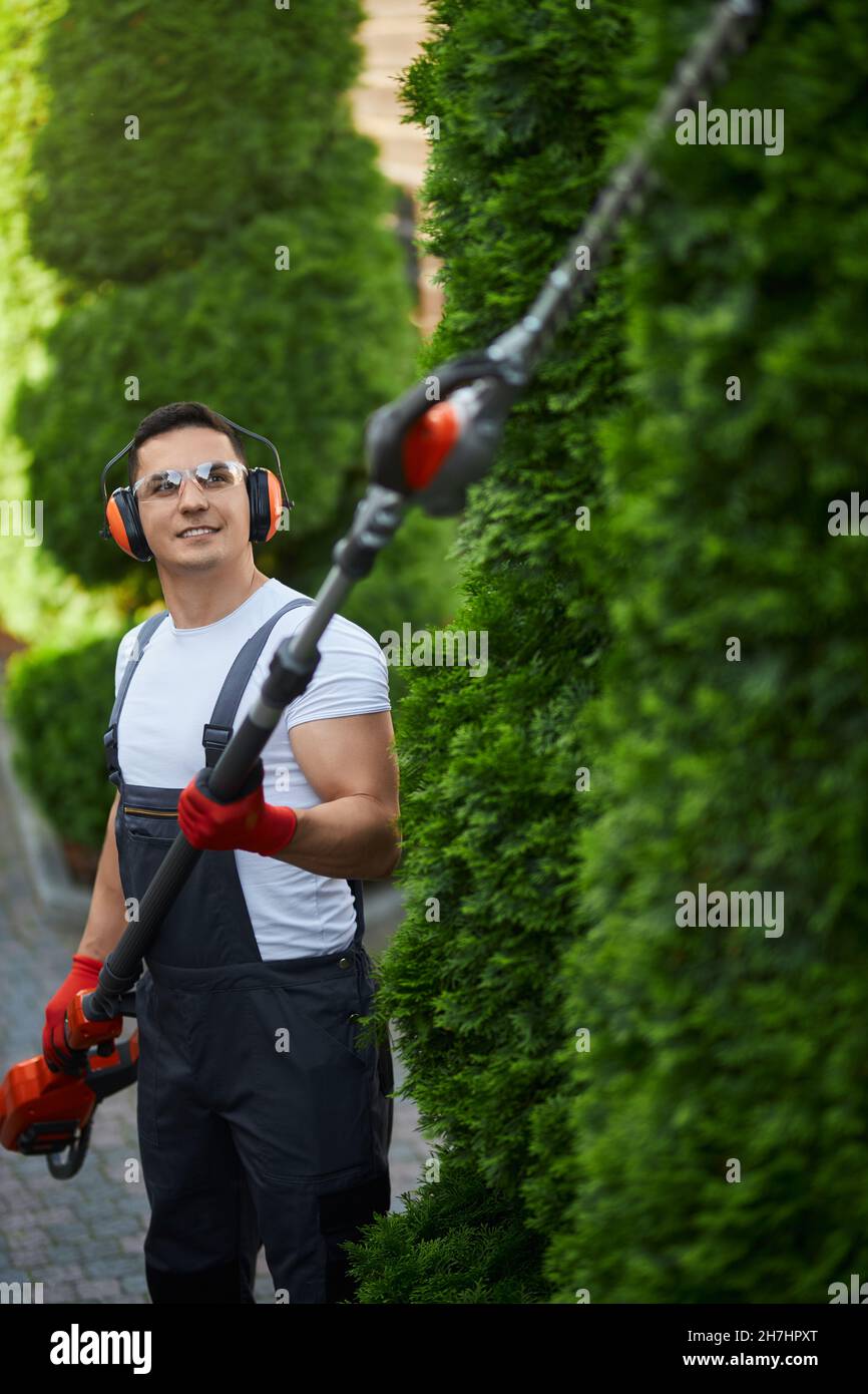 Jardinier mâle compétent utilisant une taille-haie à essence pour façonner les buissons à l'extérieur.Jeune homme caucasien portant des lunettes de protection, des écouteurs et des gants lorsqu'il travaille dans le jardin. Banque D'Images