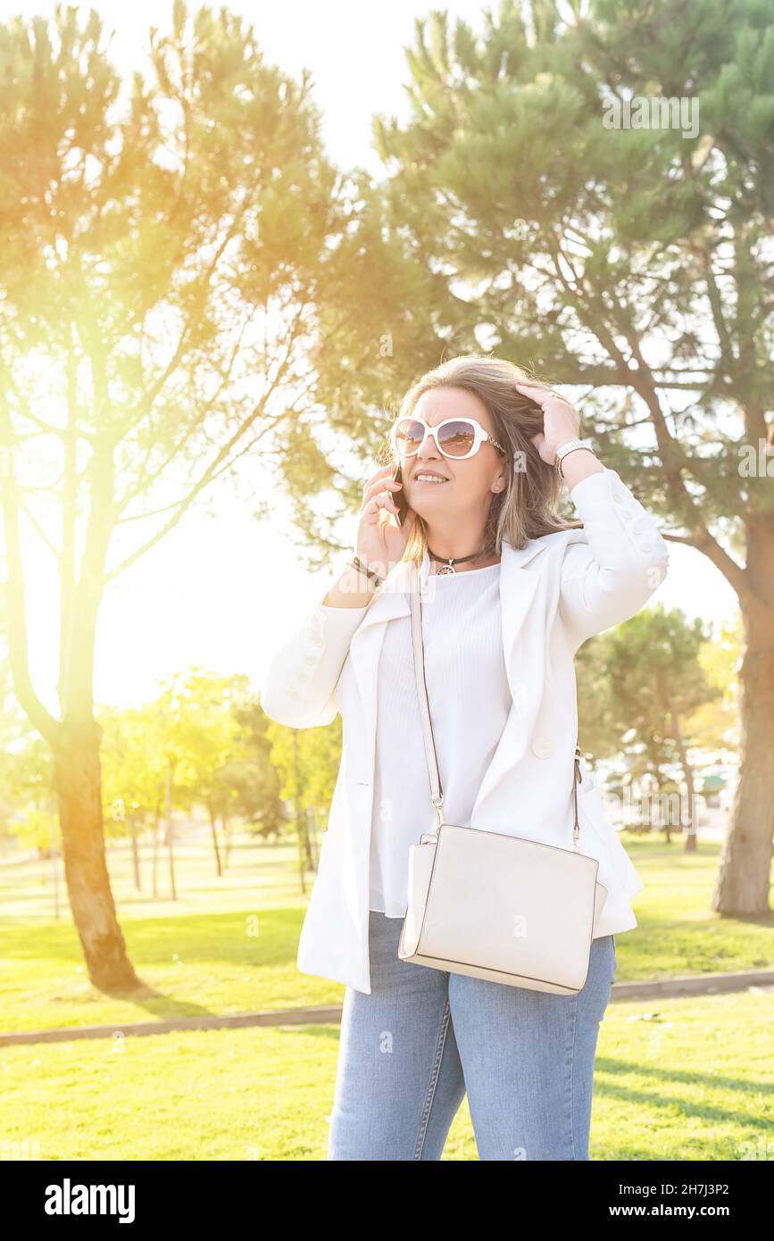 Femme d'âge moyen de race blanche debout dans un parc souriant et parlant sur son téléphone cellulaire tout en touchant ses cheveux Banque D'Images