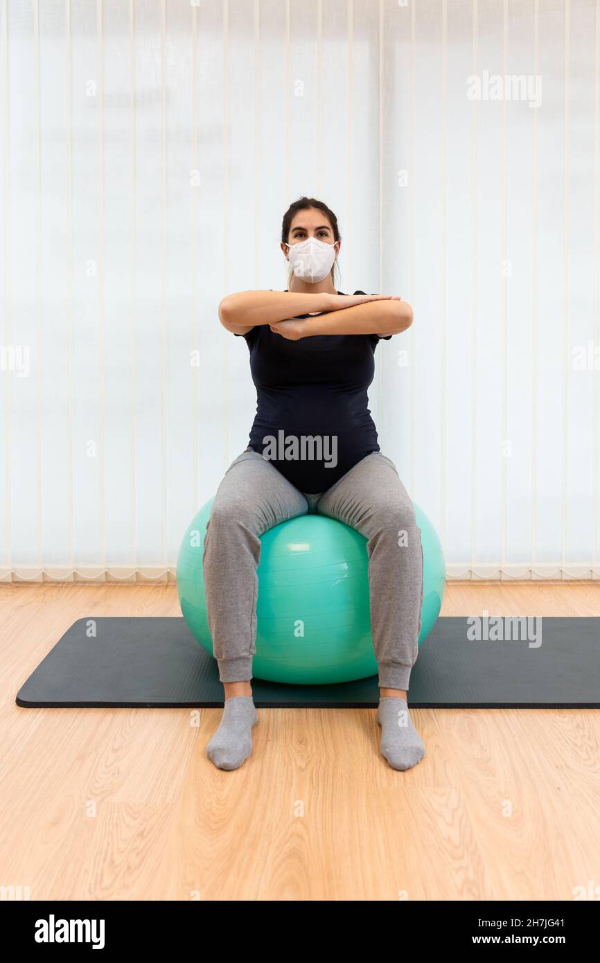 femme enceinte portant un masque facial faisant des exercices pilates assis sur une balle de fitness avec ses bras devant Banque D'Images