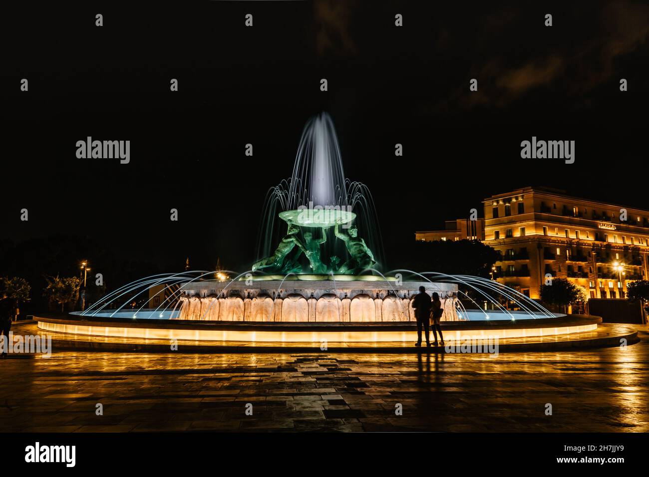 Célèbre fontaine illuminée de Tritons la nuit, trois sculptures mythologiques de Tritons en bronze tenant un immense bassin, point de repère moderne de la Valette. Banque D'Images