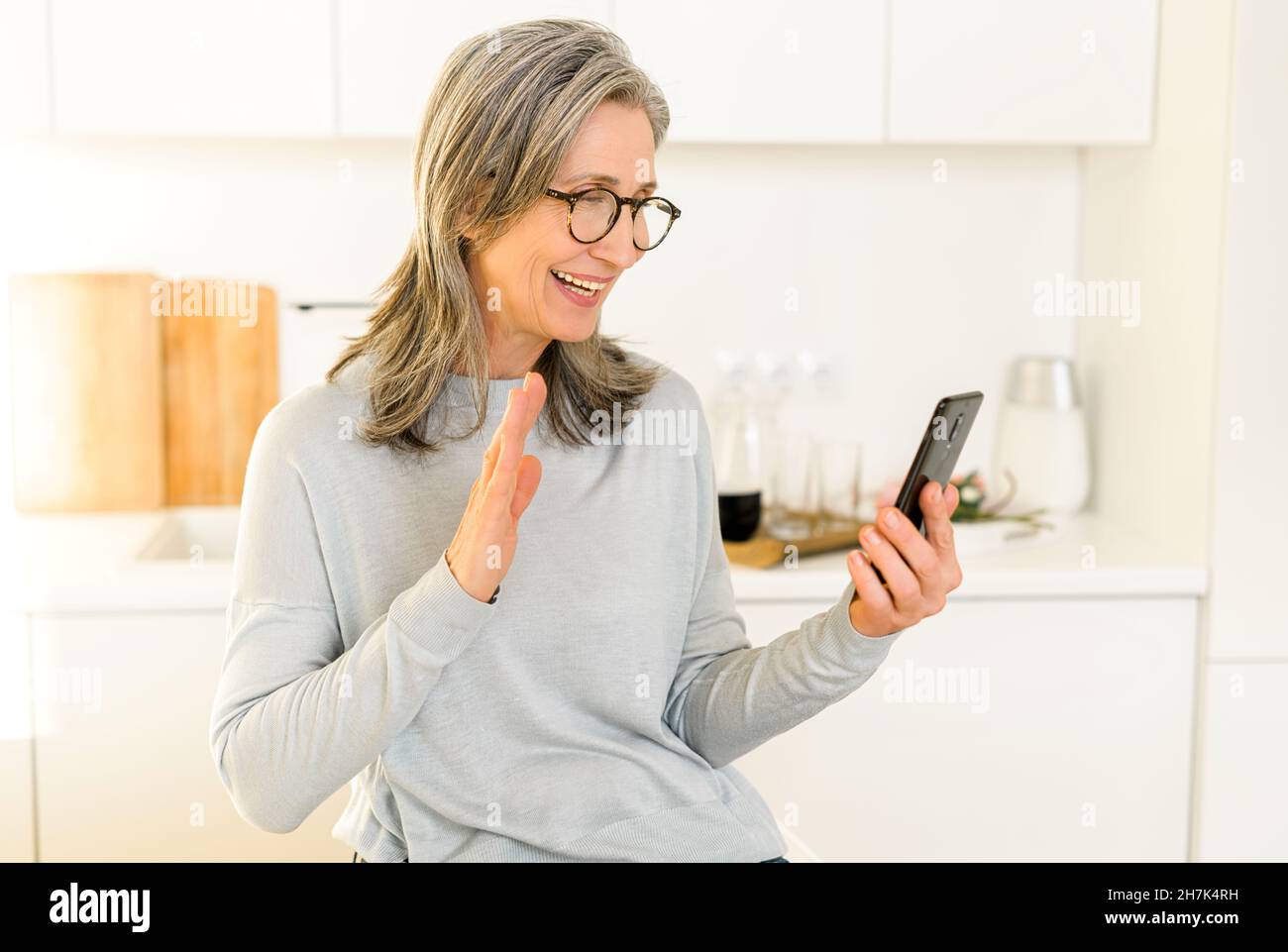 Heureuse femme mûre souriante assise dans la cuisine, se détendant et parlant à son ami ou sa famille via la connexion vidéo, femme sénior aux cheveux gris tenant un téléphone portable et se portant la main Banque D'Images