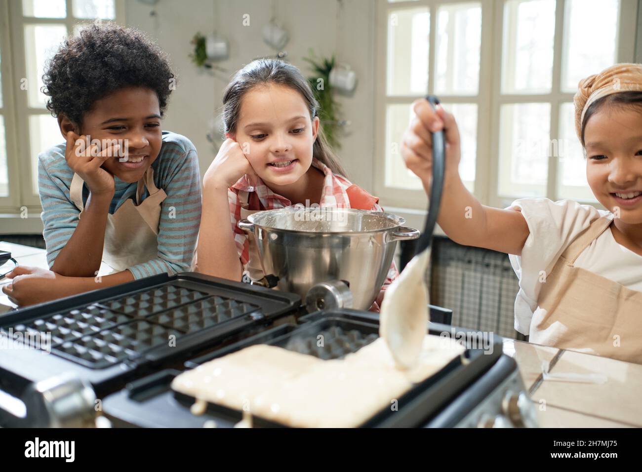 Fille mettant de la pâte sur l'icône en forme de gaufres et apprenant à cuire des gaufres avec ses amis dans la cuisine Banque D'Images