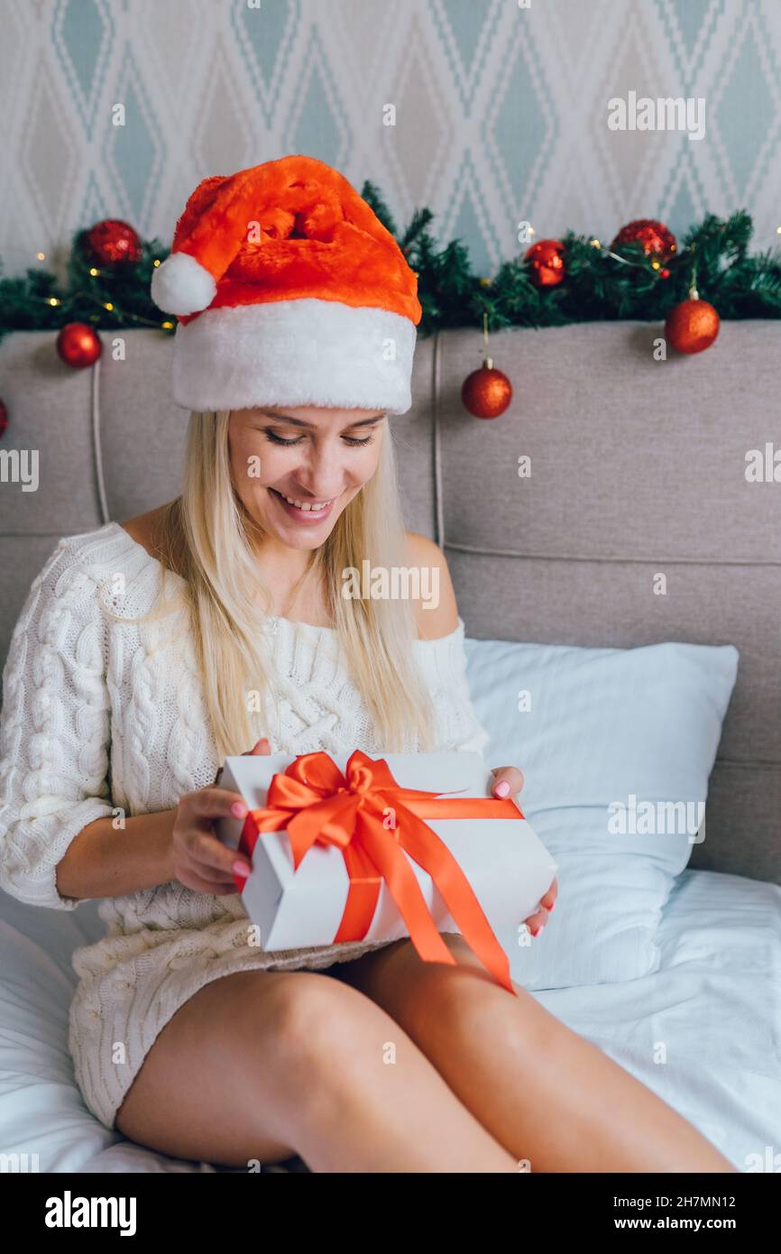 Belle jeune femme positive portant un chandail blanc tricoté et un chapeau de père noël tenant une boîte cadeau enveloppée dans un ruban rouge assis sur le lit dans la chambre à la maison. Banque D'Images