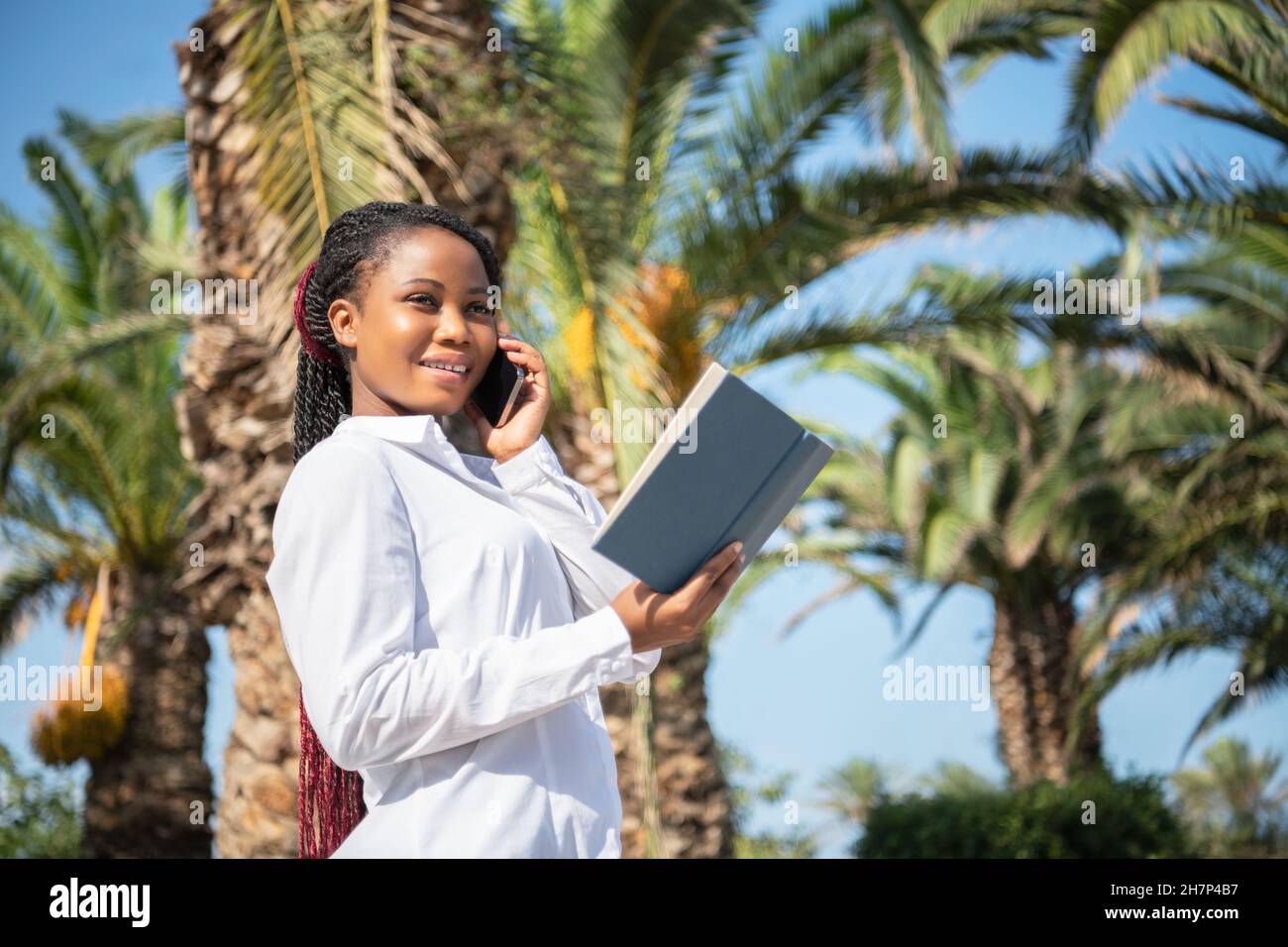 Femme africaine tenant un livre tout en parlant avec le mobile en plein air Banque D'Images