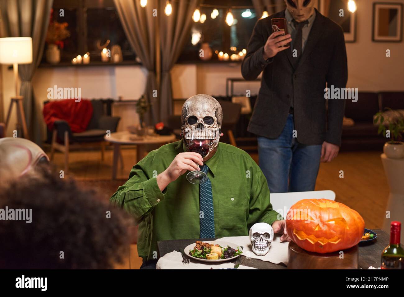 Portrait d'un homme dans un masque effrayant buvant du vin rouge tout en étant assis à la table et célébrant la fête d'Halloween avec ses amis Banque D'Images