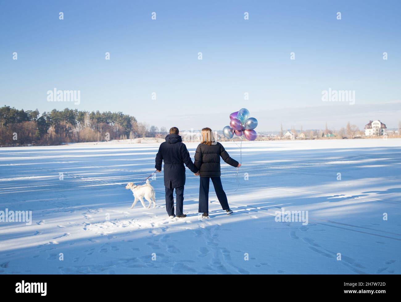 amoureux méconnaissables homme et femme dans des vêtements chauds, tenant les mains, marcher avec un chien blanc dans la neige hiver ensoleillé jour. sentiments tendres, humeur festive. Banque D'Images