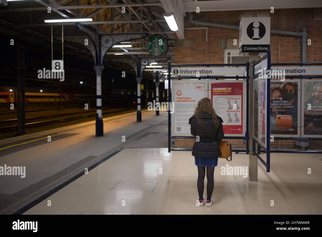 Doncaster, Royaume-Uni, 22 mai 2021: Une femme porte un manteau d'hiver la nuit, tout en lisant les informations sur une plate-forme propre de la gare de doncaster Banque D'Images