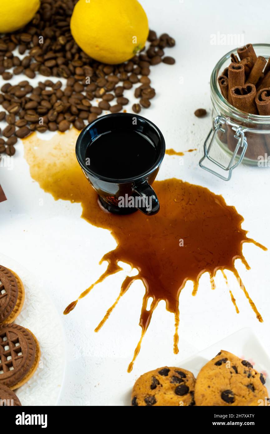 Café renversé sur une table blanche à l'aide d'une tasse noire.Grains de café, cannelle et biscuits.Petit déjeuner dans la cuisine. Banque D'Images