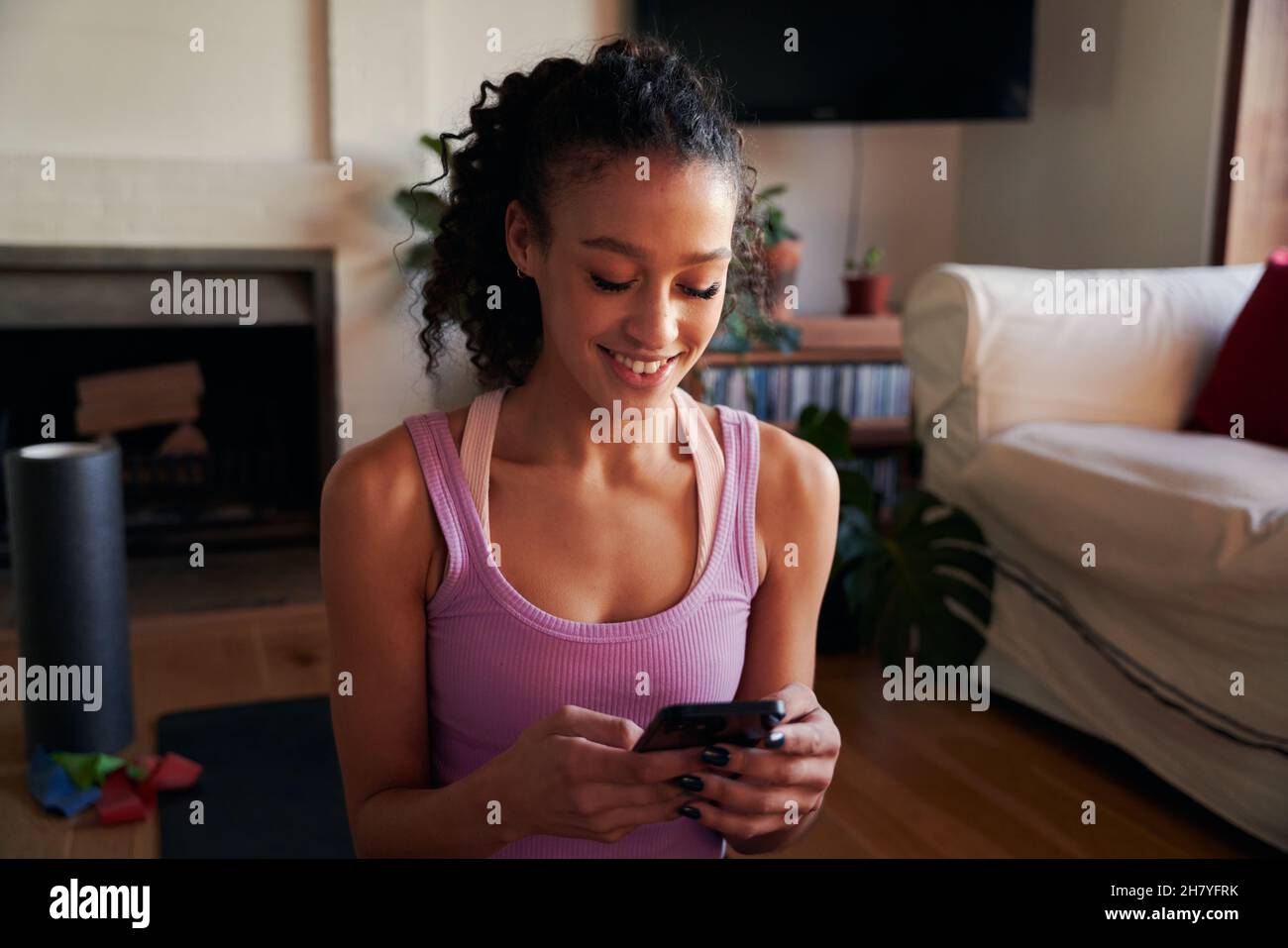 Jeune femme adulte multiculturelle souriant et envoyant des SMS sur un tapis de yoga dans le salon moderne de la maison Banque D'Images