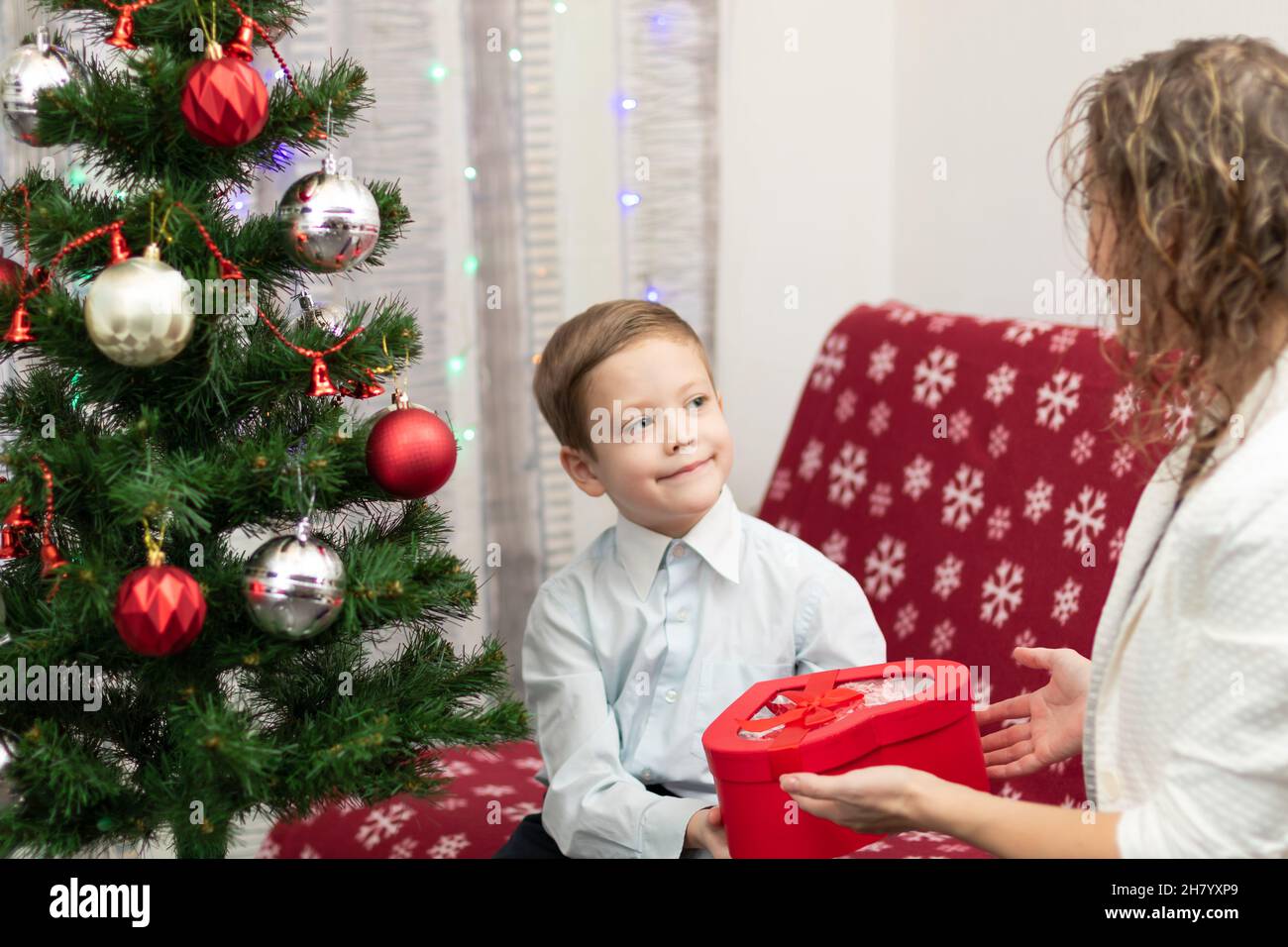 Un garçon de sept ans dans une chemise élégante offre à sa mère un cadeau dans une boîte pour le nouvel an sur fond d'un arbre de Noël à la maison.Sélectif FO Banque D'Images