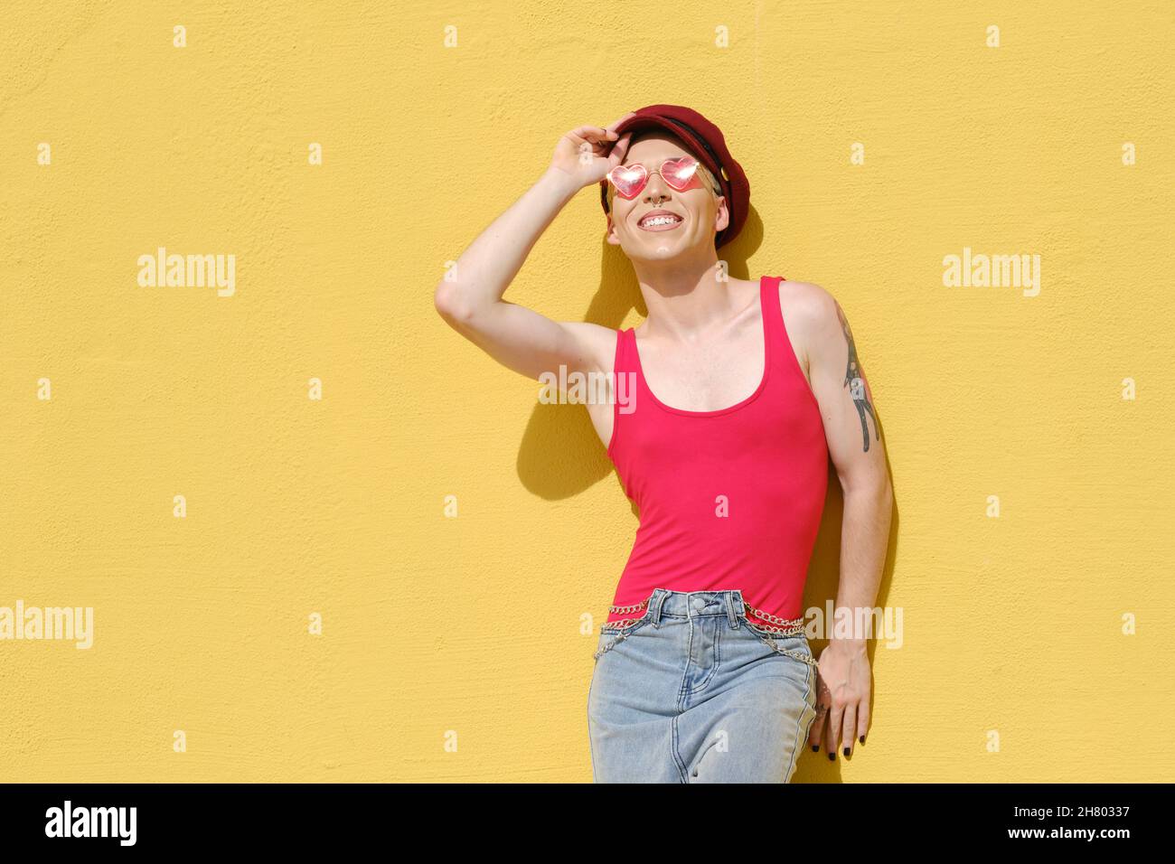 Jeune modèle non binaire souriant tout en posant à l'extérieur appuyé contre un mur jaune. Banque D'Images