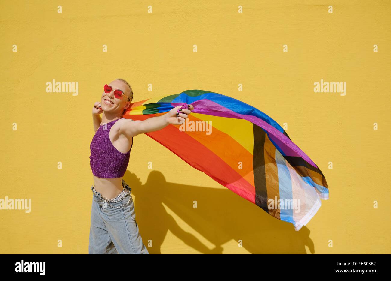 Jeune personne élégante non binaire portant le drapeau lgbt arc-en-ciel contre un mur jaune. Banque D'Images