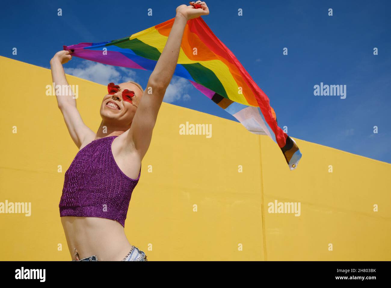 Jeune personne non binaire qui a l'air heureux et excité tout en agitant un drapeau arc-en-ciel à l'extérieur. Banque D'Images