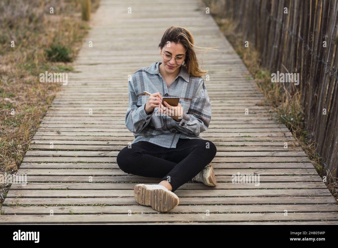 Tout le corps de la jeune femme pensive en vêtements décontractés et lunettes de vue prenant des notes et regardant loin tout en étant assis sur un chemin de bois menant à la mer sous le nuage Banque D'Images