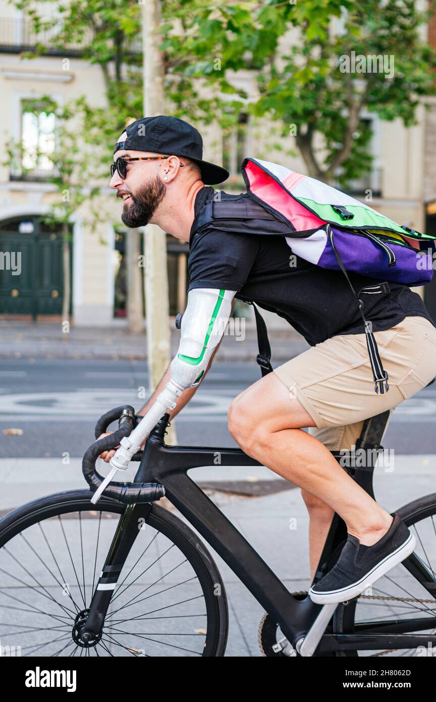 Vue latérale de l'hipster mâle barbu avec bras artificiel à vélo le long de la rue en été dans la ville Banque D'Images