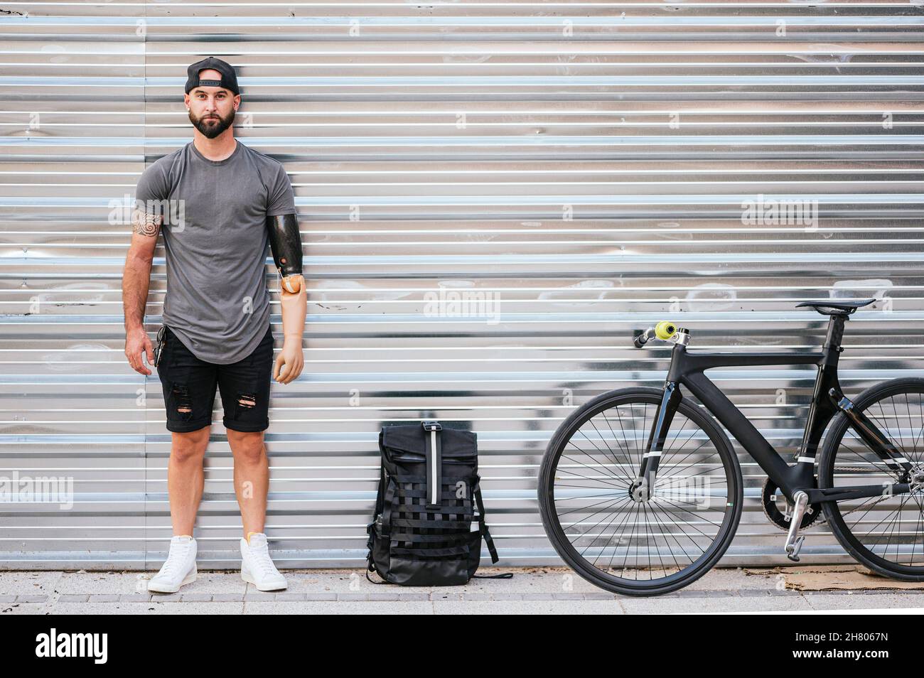 Culotte taille basse homme confiante avec bras artificiel à proximité d'un sac à dos et d'un vélo en ville et regardant l'appareil photo contre un mur en métal Banque D'Images