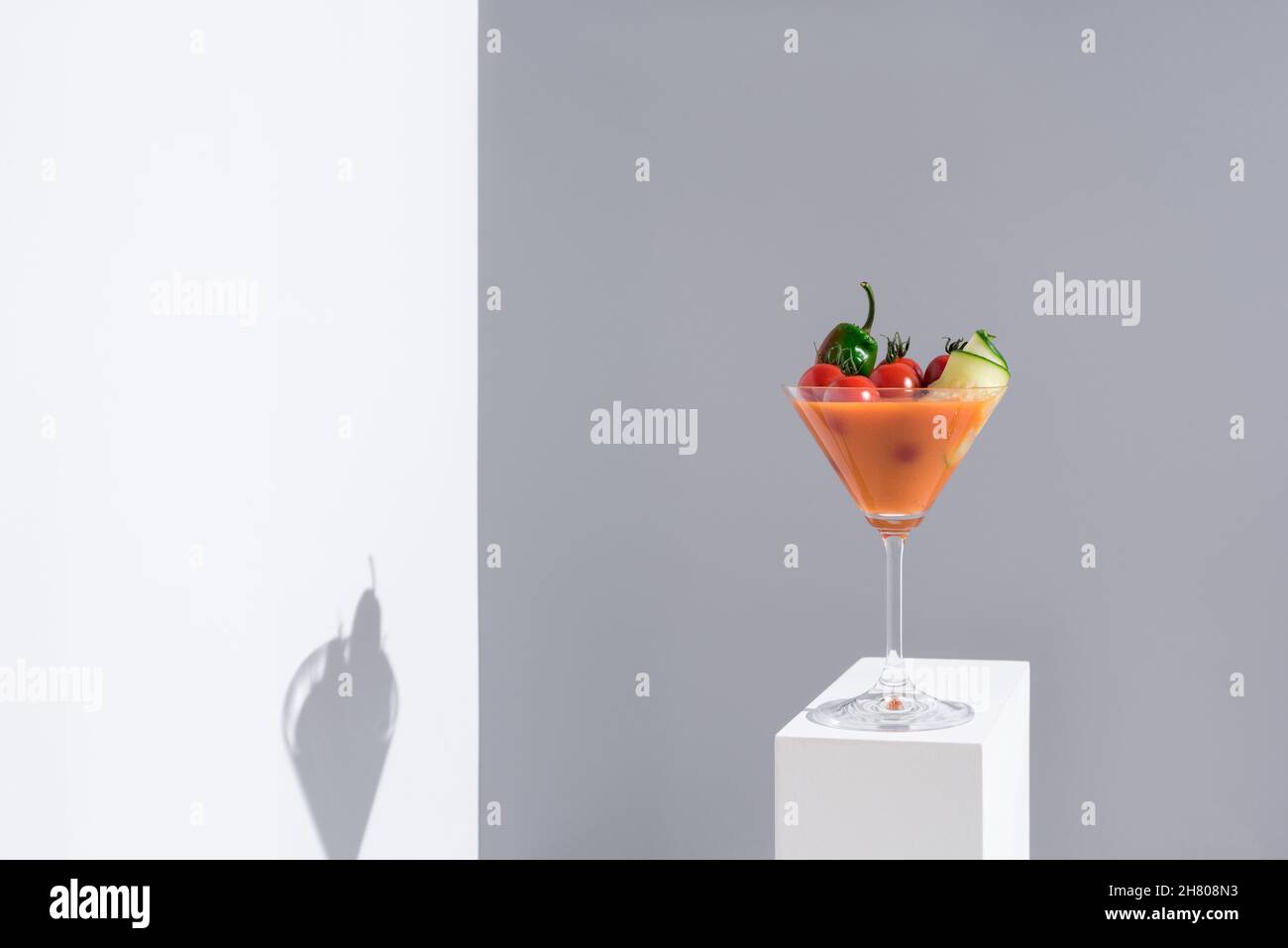 Soupe fraîche de gazpacho avec tomates cerises et concombres servis dans un verre à cocktail placé sur un support blanc sur fond gris Banque D'Images