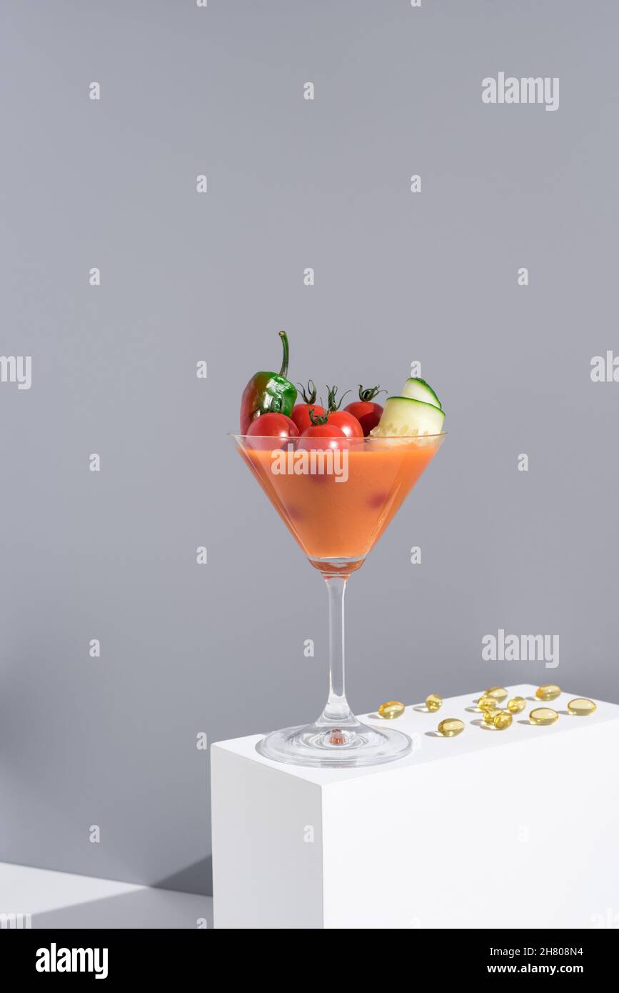 Soupe fraîche de gazpacho avec tomates cerises et concombres servis dans un verre à cocktail placé sur un support blanc sur fond gris Banque D'Images