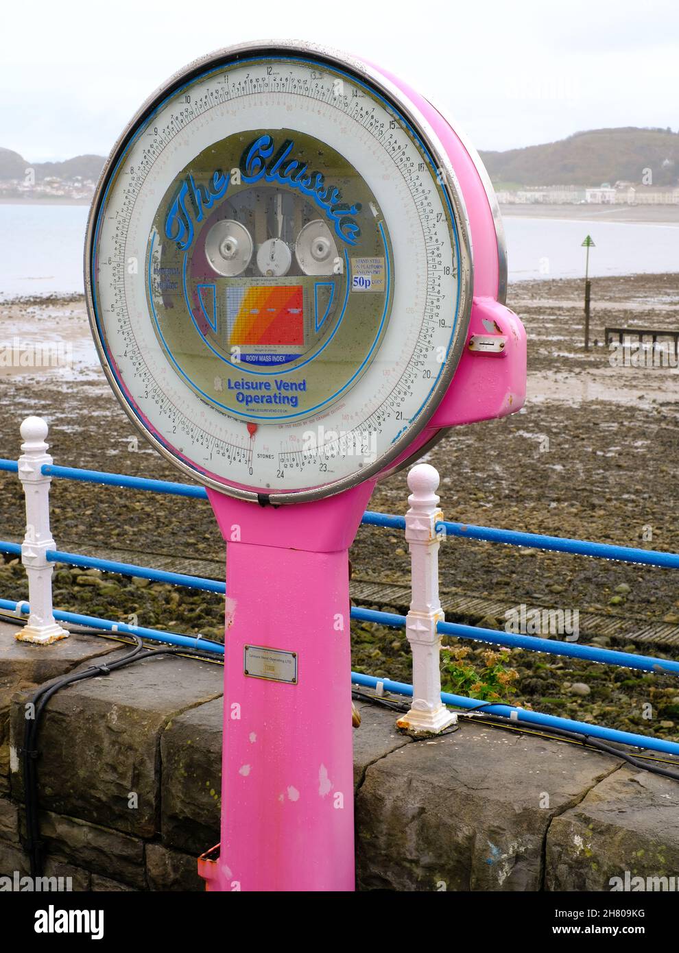 Une machine de pesage traditionnelle rose vif se trouve sur la jetée de Llandudno et contraste avec les tons doux de la plage rocheuse au-delà. Banque D'Images