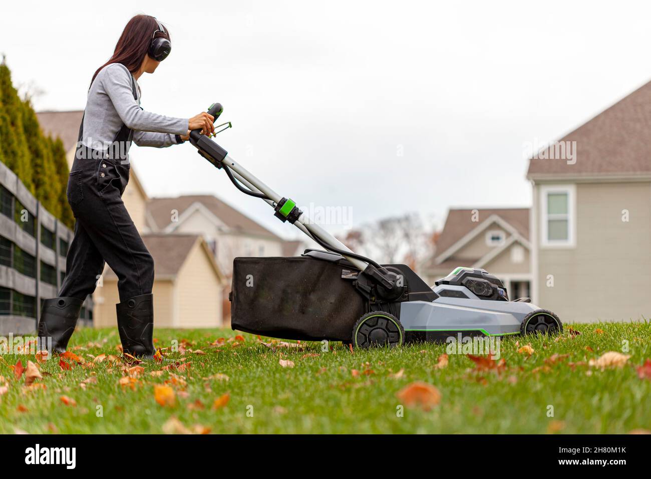 une jeune femme du caucase portant une combinaison et des inhibiteurs de bruit fait fonctionner un moteur à gazon sur l'herbe à la fin de l'automne.Il y a des feuilles tombées et un suburbain n Banque D'Images